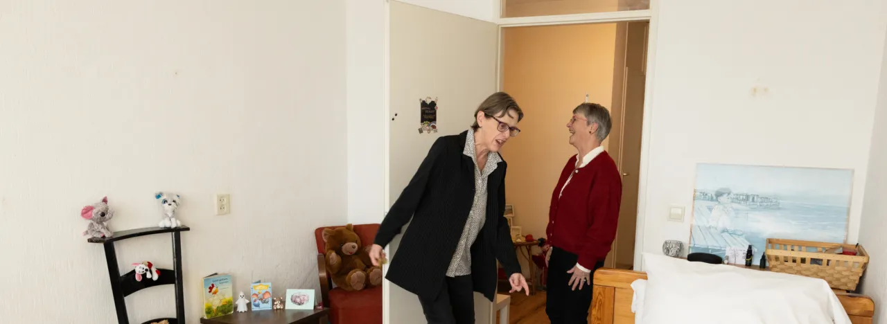 Twee vrouwen in een slaapkamer. Er staat een bed met een tovenaarshoed er op. De ene vrouw moet lachen